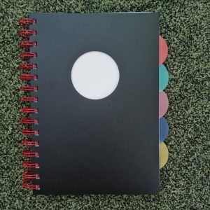 black notebook with red ringbinding and coloured page dividers showing on the right side, lying on a green carpet